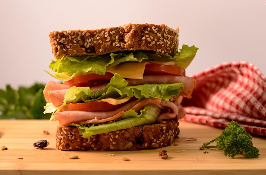 A whole-grain sandwich with sesame seeds, filled with lettuce, tomato, cheese and ham, on a wooden board.
