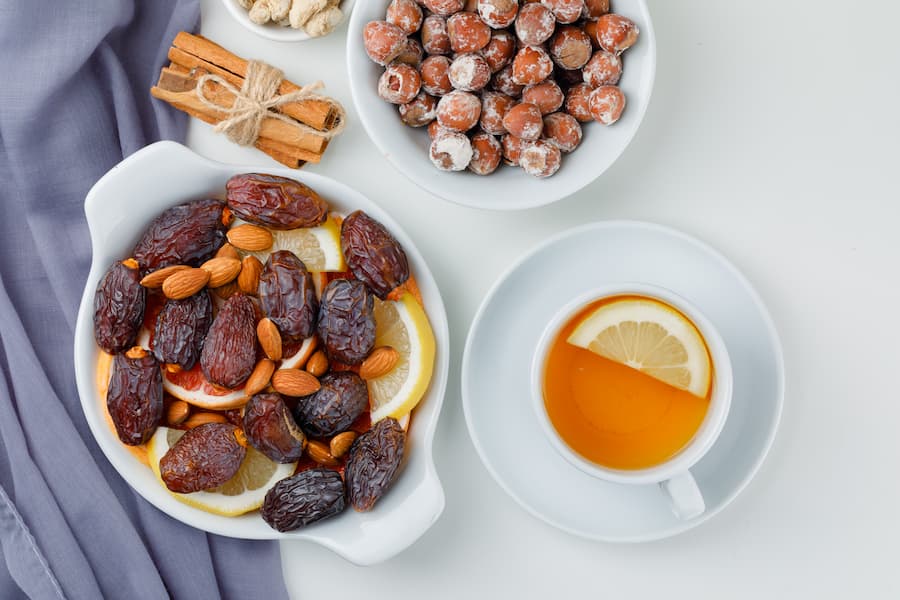 Dates and almonds on a white dish with citrus slices, beside a bowl of hazelnuts, cinnamon sticks and a cup of lemon tea.