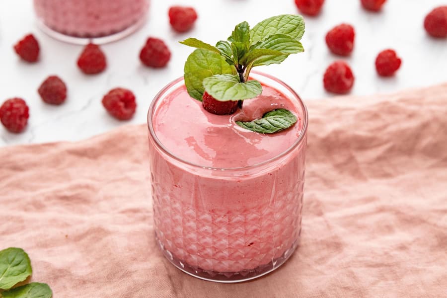 A glass of pink fruit smoothie garnished with fresh mint leaves and a raspberry, set on a pink cloth with raspberries scattered around it.