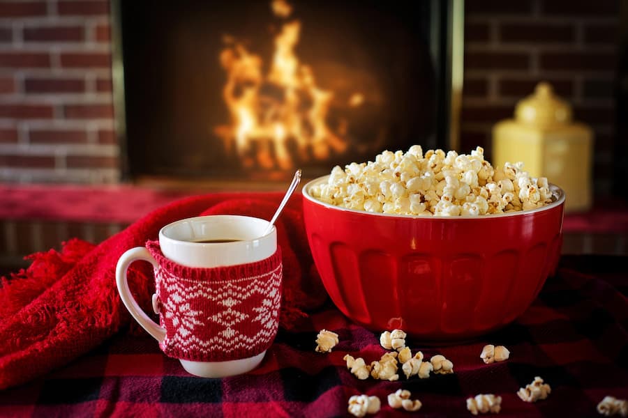 Red bowl of popcorn next to a mug of coffee in a knitted sleeve by a fireplace
