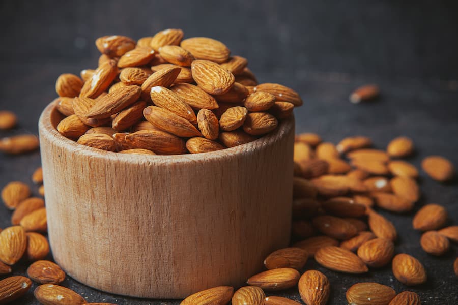 Wooden bowl of almonds with scattered almonds on a dark surface